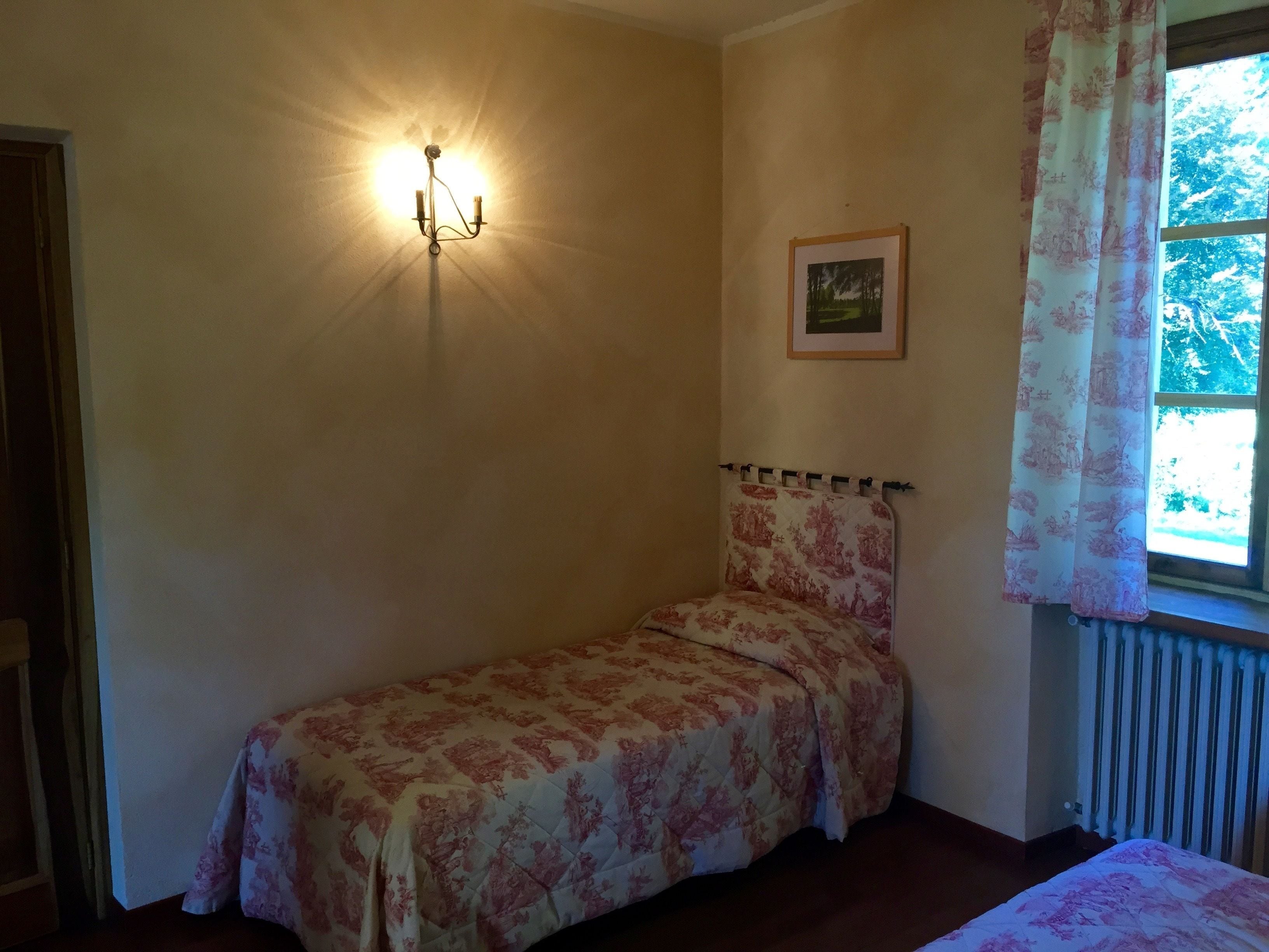 Une petite chambre avec un lit simple, une couvre-lit à motifs floraux, une lampe fixée au mur, un tableau encadré et une fenêtre avec des rideaux.