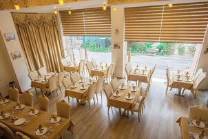 Un ristorante con diversi tavoli e sedie, ampie finestre con tende e vista sull'esterno.