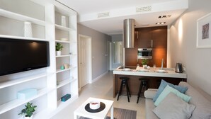 Una sala de estar moderna con un televisor de pantalla plana, estanterías blancas, una mesita pequeña blanca y un sofá gris con cojines estampados.