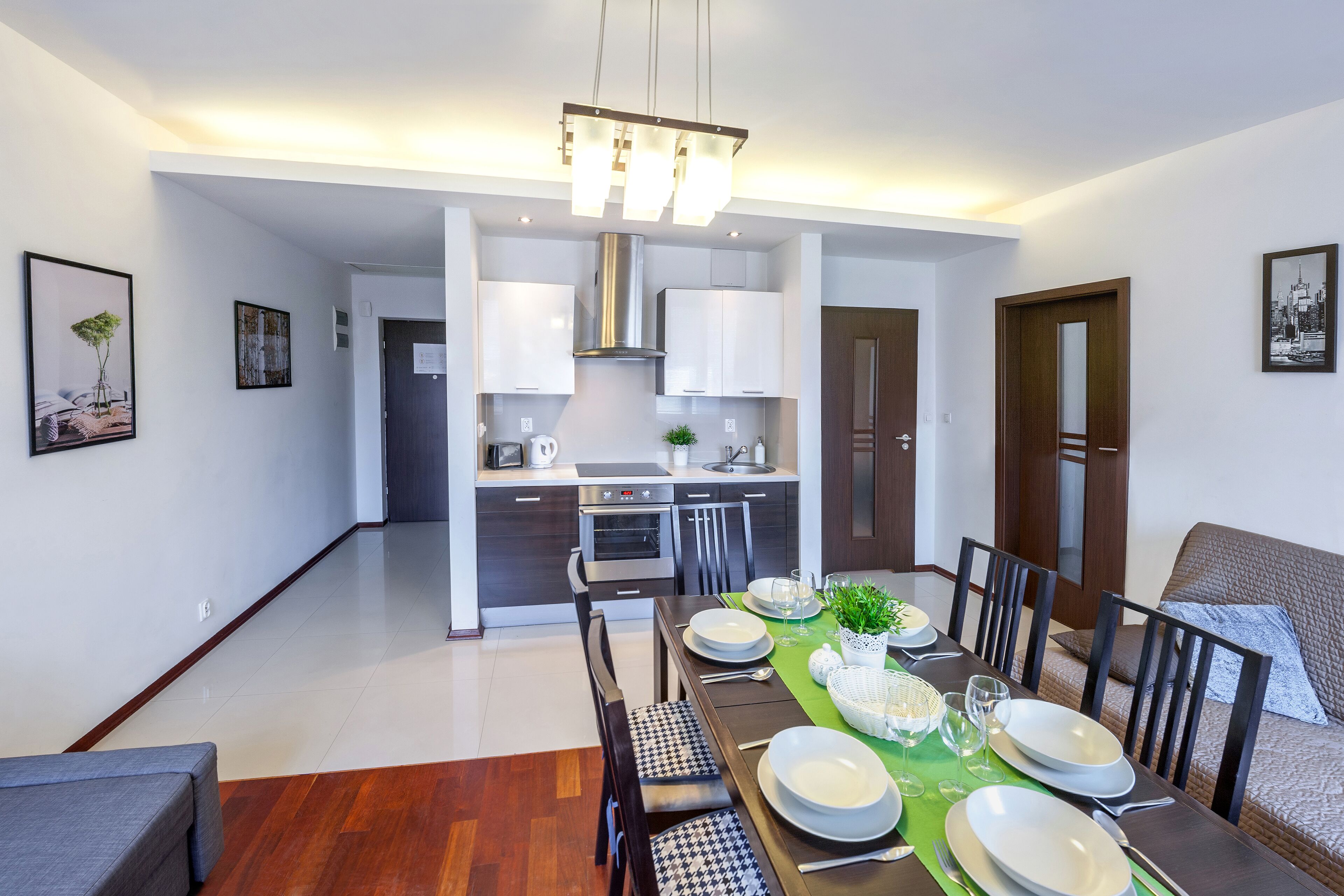 Uma sala de jantar moderna com uma mesa comprida posta para quatro pessoas, cozinha ao fundo, e um espaço de estar acolhedor com um sofá e poltronas.
