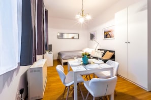 Quarto moderno e bem iluminado com uma mesa de jantar posta para dois, uma cama com cobertor cinza, uma mesa de cabeceira com abajur e uma grande janela com cortinas.