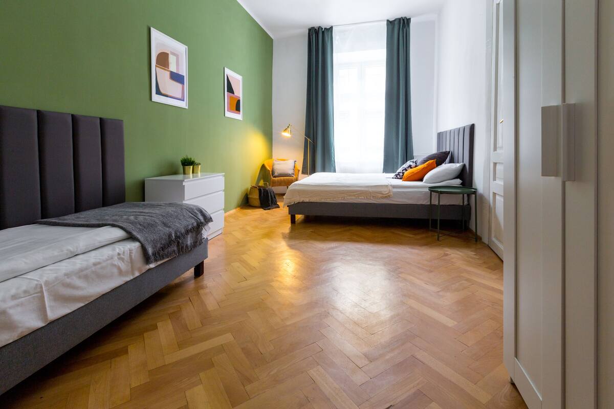 A modern hotel room with two beds, a wooden floor, a green wall, and a large window with curtains.