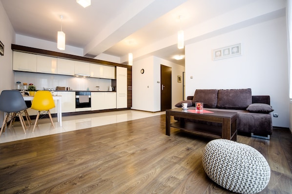 A modern living room with a wooden floor, a sofa, a coffee table, and a dining area with chairs and a kitchen in the background.