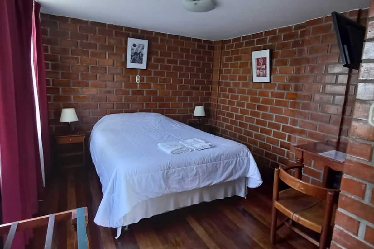Una habitación con pared de ladrillo, una cama con cubrecama blanco, dos mesitas de noche con lámparas y una silla de madera.