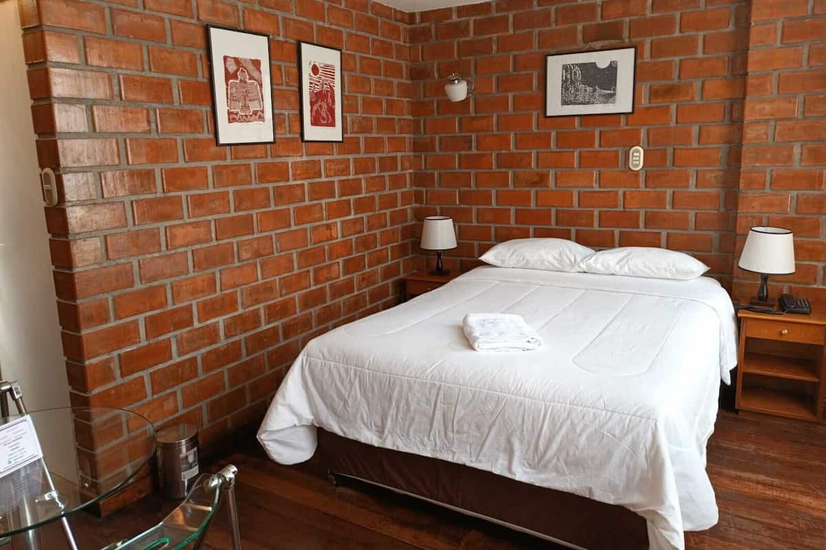 Una habitación de hotel con pared de ladrillo, una cama con ropa de cama blanca, dos mesitas de noche con lámparas y cuadros en la pared.
