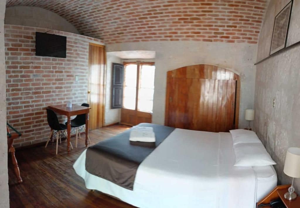 Habitación de hotel con pared de ladrillo, cabecera de madera, cama con ropa blanca, escritorio con silla y ventana con cortinas.