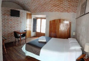 A hotel room with a brick wall, a wooden headboard, a bed with white linens, a desk with a chair, and a window with curtains. - La Casa de Margott (Arequipa)