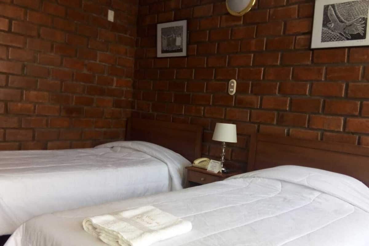 Una habitación con dos camas, una pared de ladrillo, una mesita de noche con una lámpara y un cuadro en la pared.