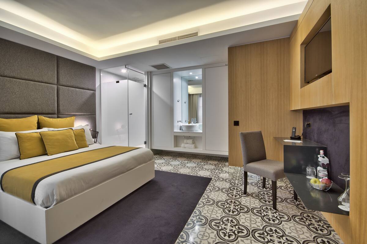 Una habitación de hotel moderna con una cama grande, zona de estar, tocador y una pequeña mesa con una botella y vasos.