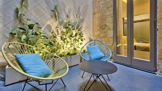 Un patio con sillas de mimbre, una mesa, una planta en maceta y un ventanal corredera que da a una habitación.