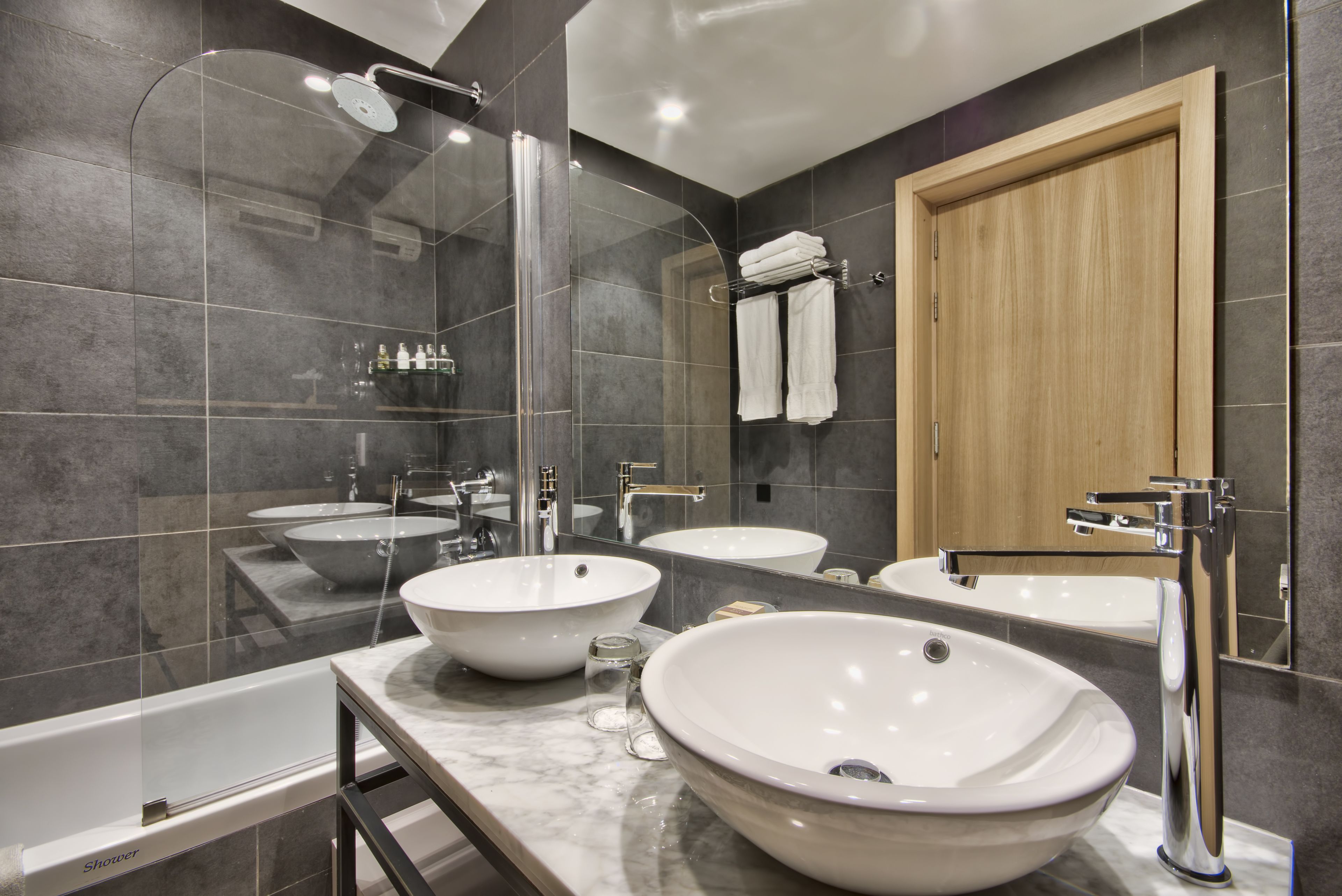 Un baño moderno con dos lavamanos blancos grandes, una ducha con cerramiento de vidrio y una puerta de madera.