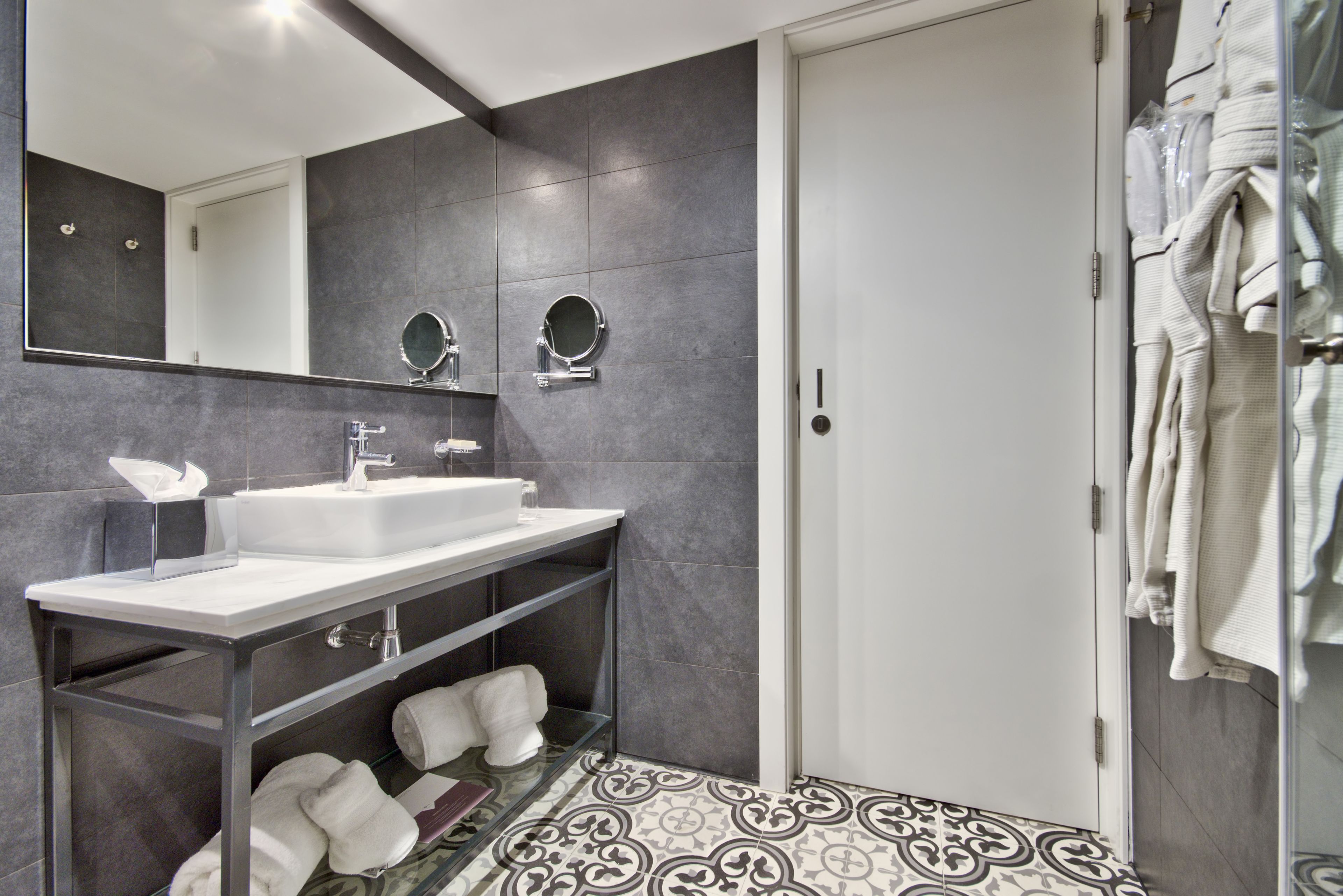 Un baño moderno con un lavabo blanco, dos espejos redondos, un perchero para toallas y baldosas de suelo estampadas.