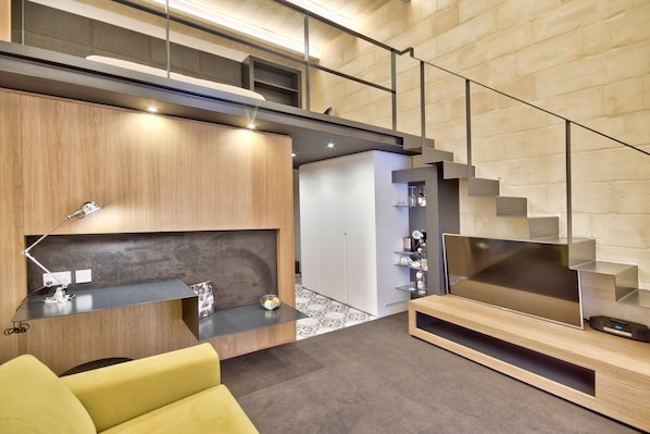 A modern living room with a sofa, a flat-screen TV, a desk with a lamp, and a staircase. - Ursulino Valletta (Valletta)
