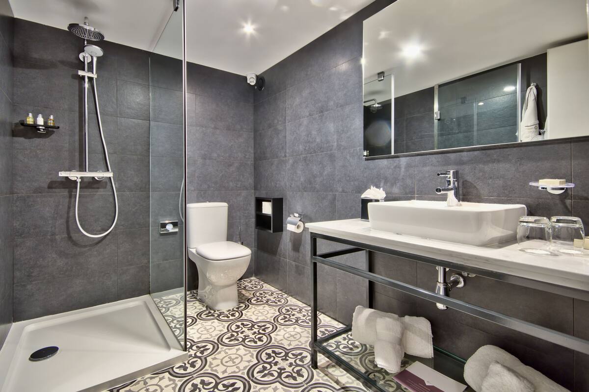 Un baño moderno con ducha, inodoro y lavabo, con baldosas oscuras y suelo estampado.