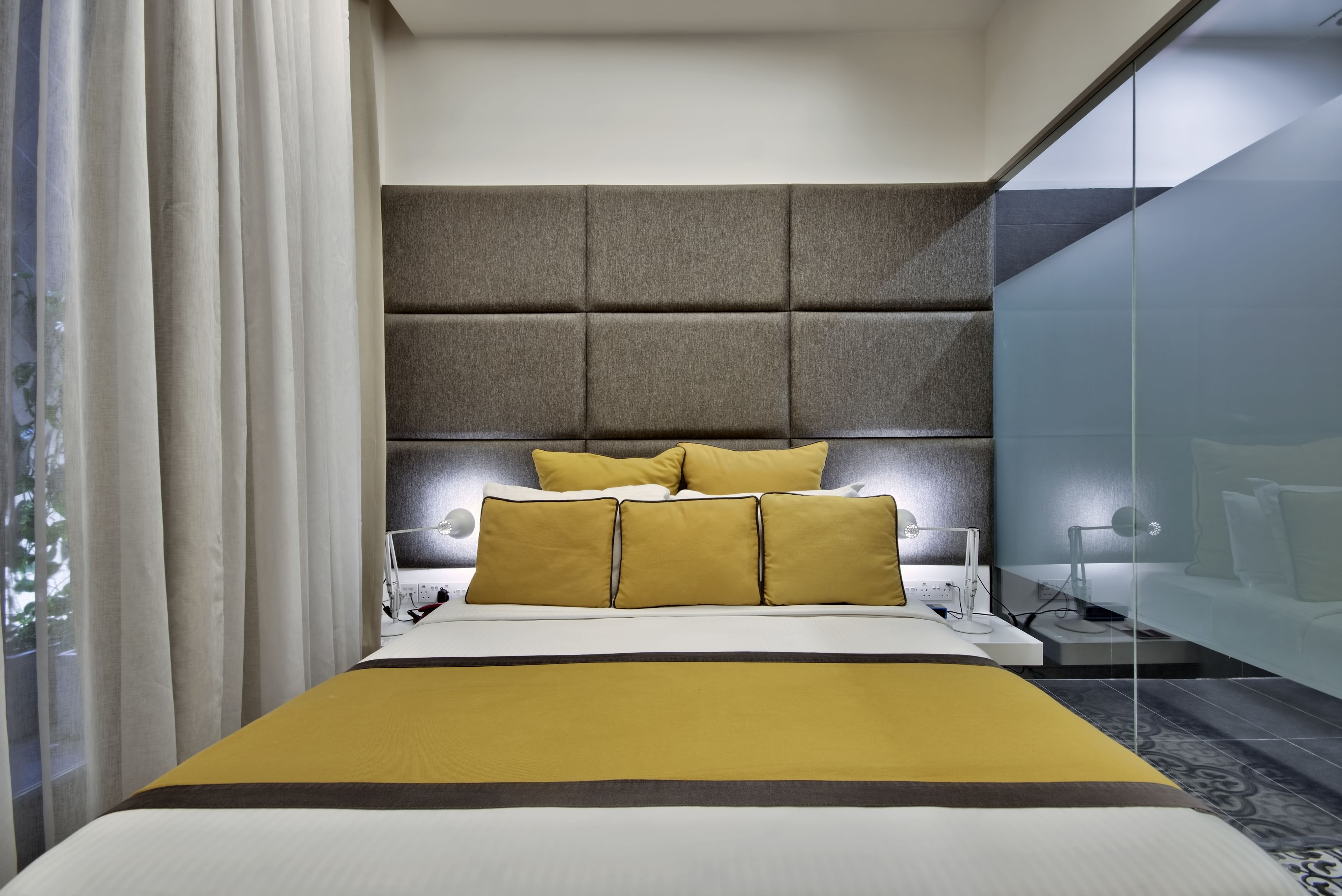 A modern hotel room with a large bed, a padded headboard, and a glass partition.