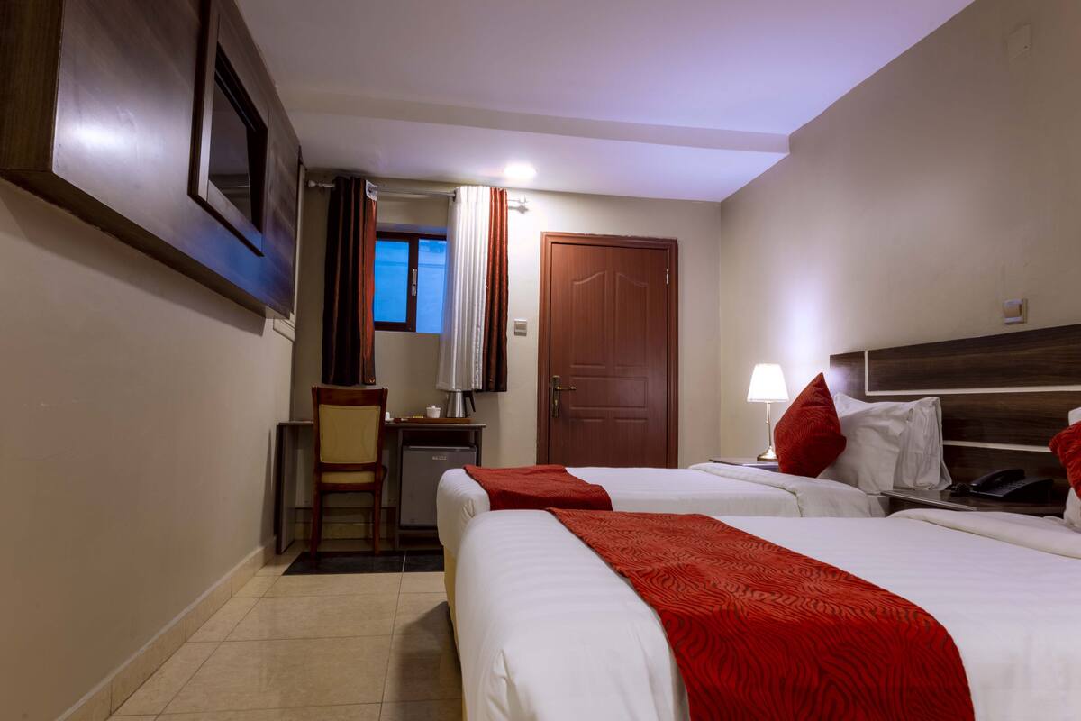 A hotel room with two beds, a desk with a lamp, a chair, and a door.