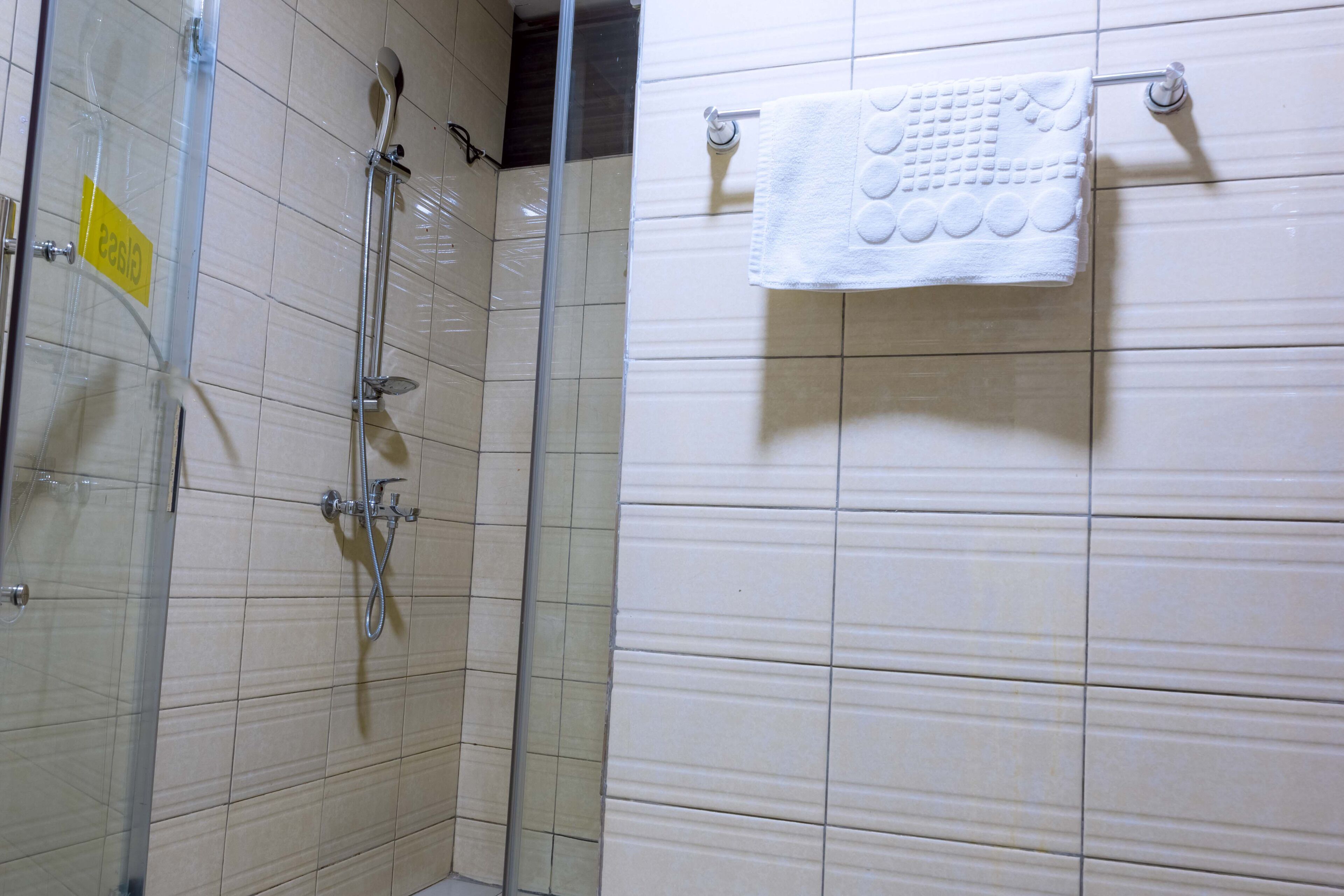 A bathroom with a glass shower enclosure, a white towel hanging on a rack, and a yellow sign on the glass door.
