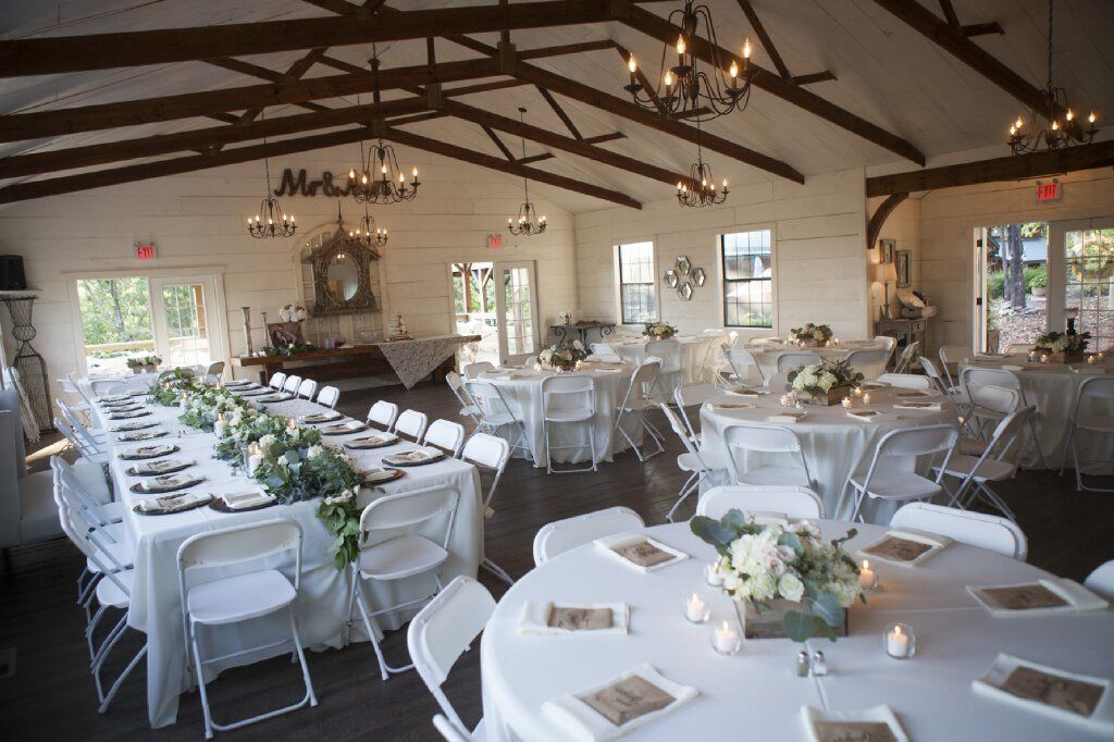 A banquet hall with multiple round tables set for dining, white chairs, chandeliers, and a rustic wooden ceiling.