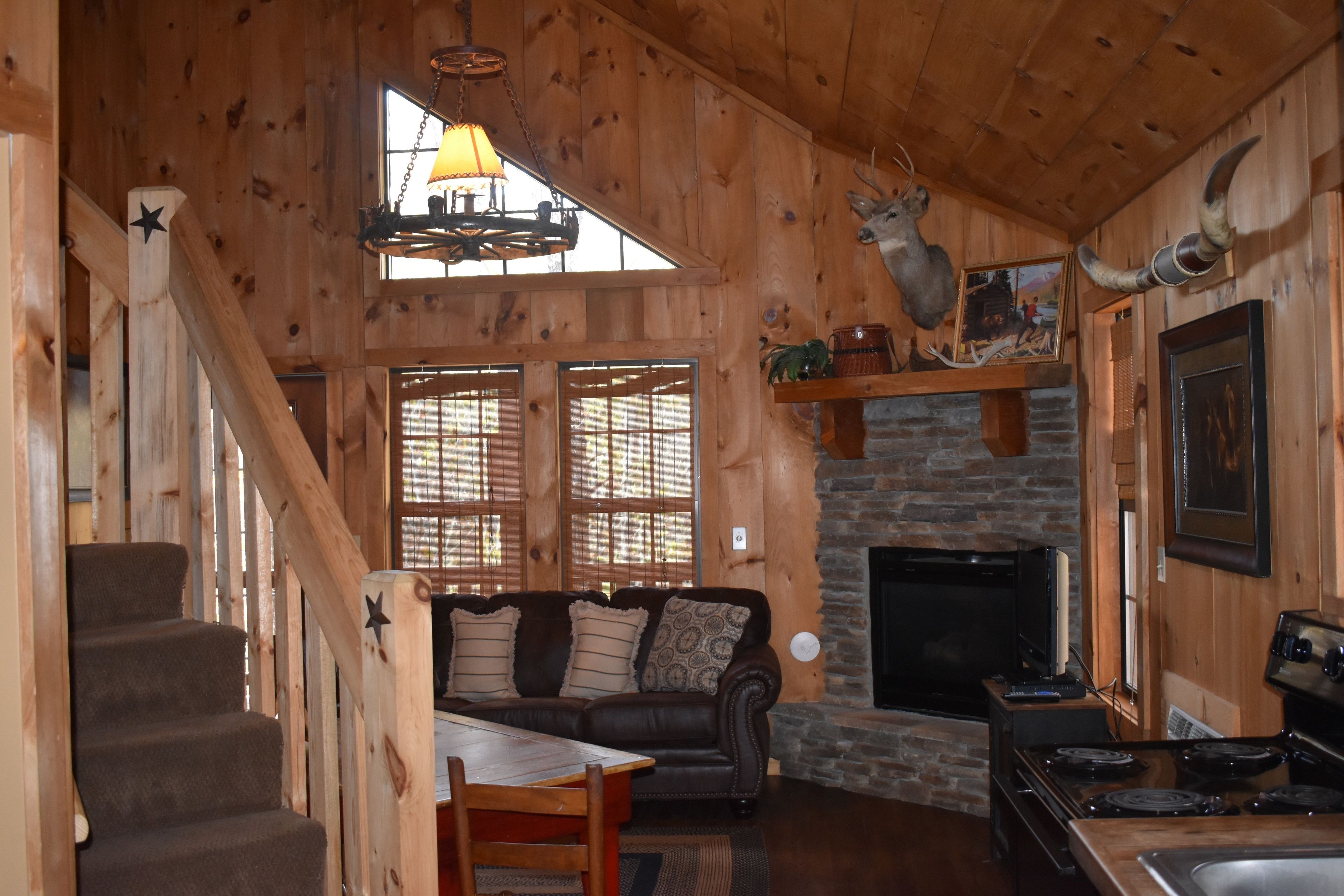 A cozy cabin interior with a stone fireplace, wooden walls, a staircase, a sofa, and a dining table.