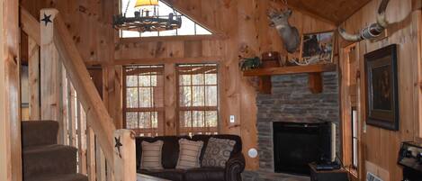 A cozy cabin interior with a stone fireplace, wooden walls, a staircase, a sofa, and a dining table.