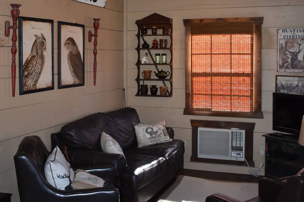 Een gezellige woonkamer met een leren bank, een kroonluchter, een bordje met de tekst 'Antiques' en een raam met jaloezieën.