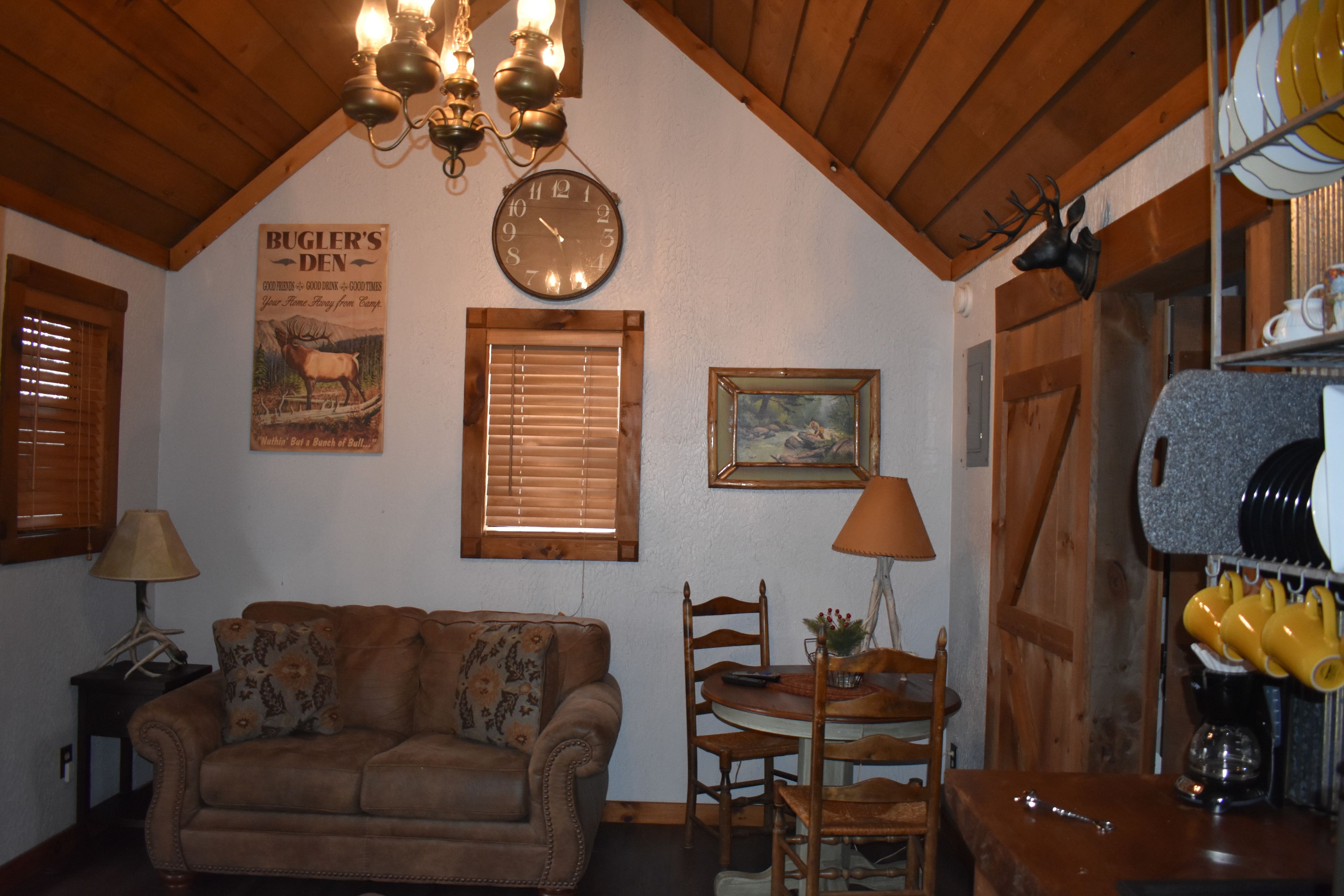 A cozy living room with a brown sofa, wooden ceiling beams, a wall clock, a framed picture, and a dining area with a table and chairs.