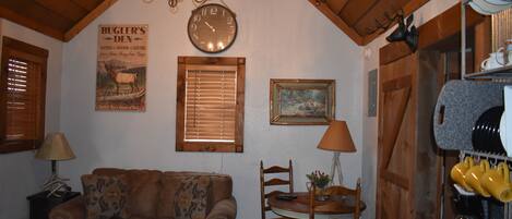 A cozy living room with a brown sofa, wooden ceiling beams, a wall clock, a framed picture, and a dining area with a table and chairs.