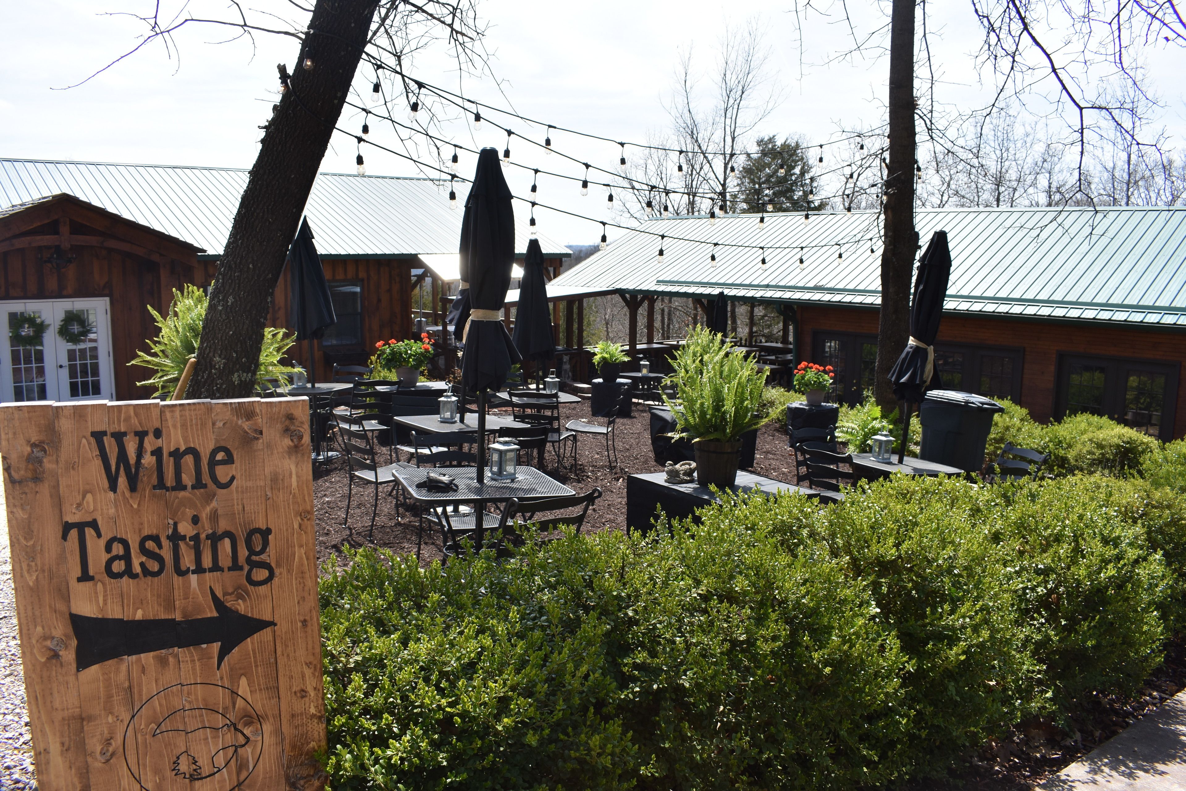 An outdoor seating area with tables and chairs, a sign indicating wine tasting, and a building with a green roof in the background.