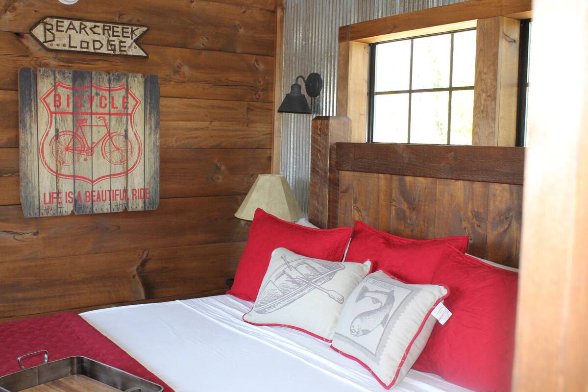 Het interieur van een houten hutje met een bed, rode en witte kussens, een bordje met de tekst 'Bear Creek Lodge' en een afbeelding van een fiets.