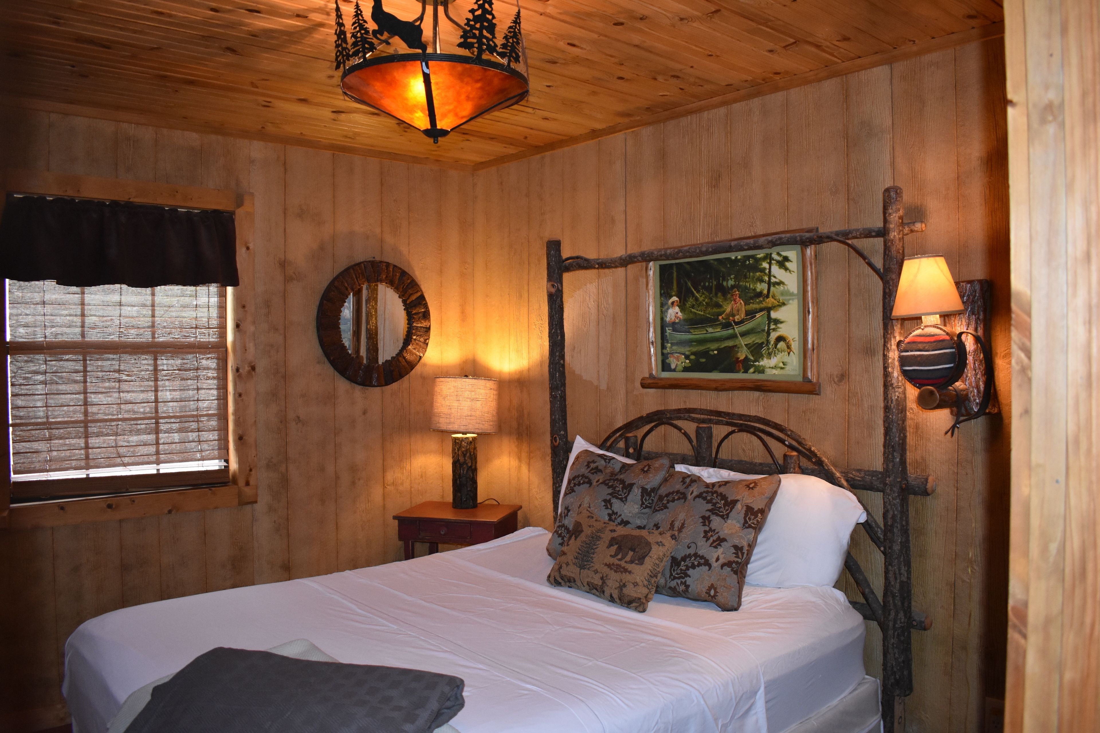 A wooden room with a bed, a window with blinds, a wall-mounted lamp, and a painting.