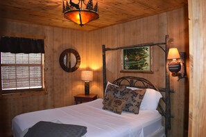 Una habitación de madera con una cama, un ventanal con persianas, una lámpara de pared y un cuadro.