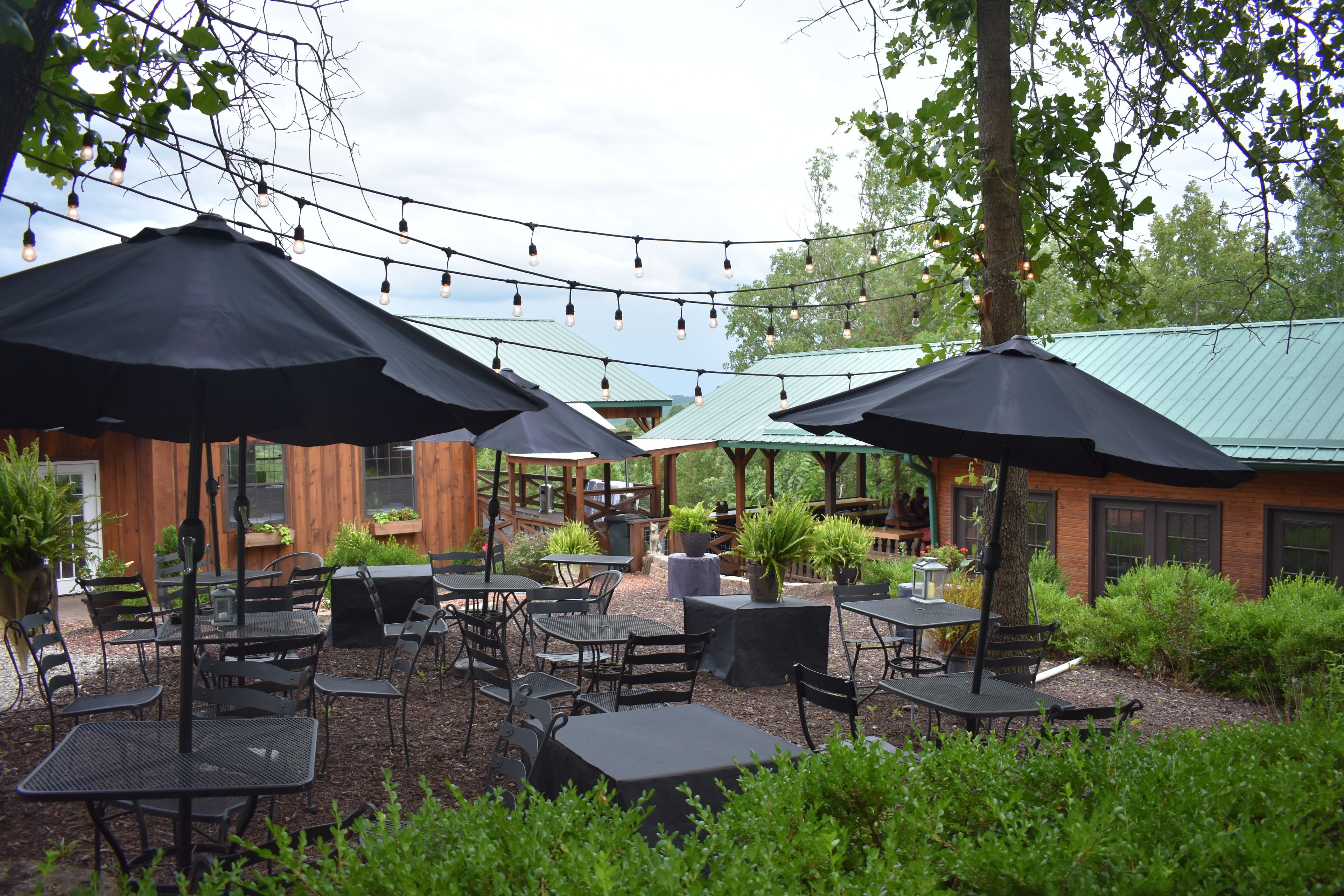 An outdoor patio area with multiple tables and chairs, string lights overhead, and a brick building in the background.