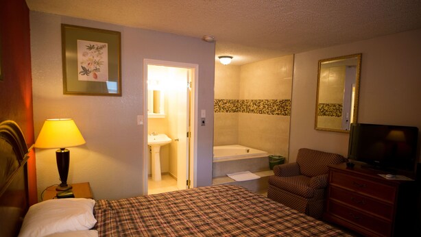 A hotel room with a bed, a chair, a dresser, a television, a lamp, a bathroom with a sink and a bathtub, and a mirror. - Scottish Inn and Suites (Bensalem)