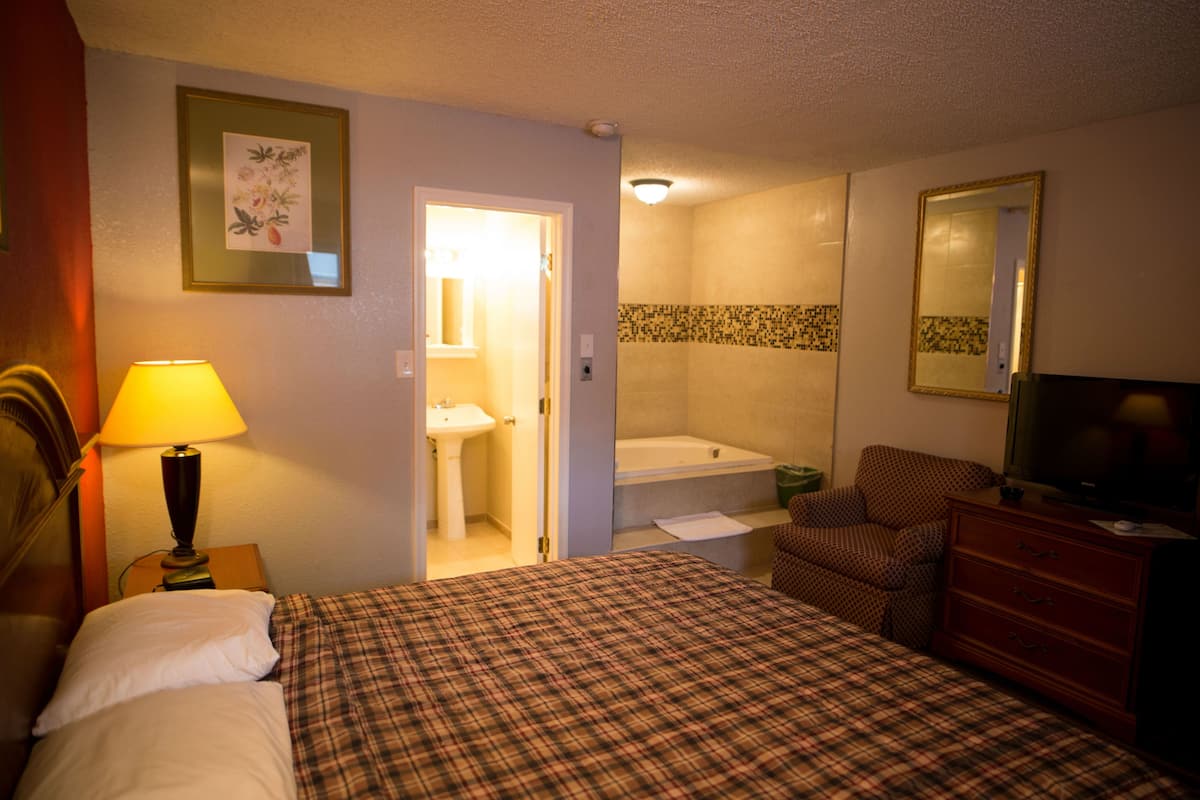 A hotel room with a bed, a chair, a dresser, a television, a lamp, a bathroom with a sink and a bathtub, and a mirror.