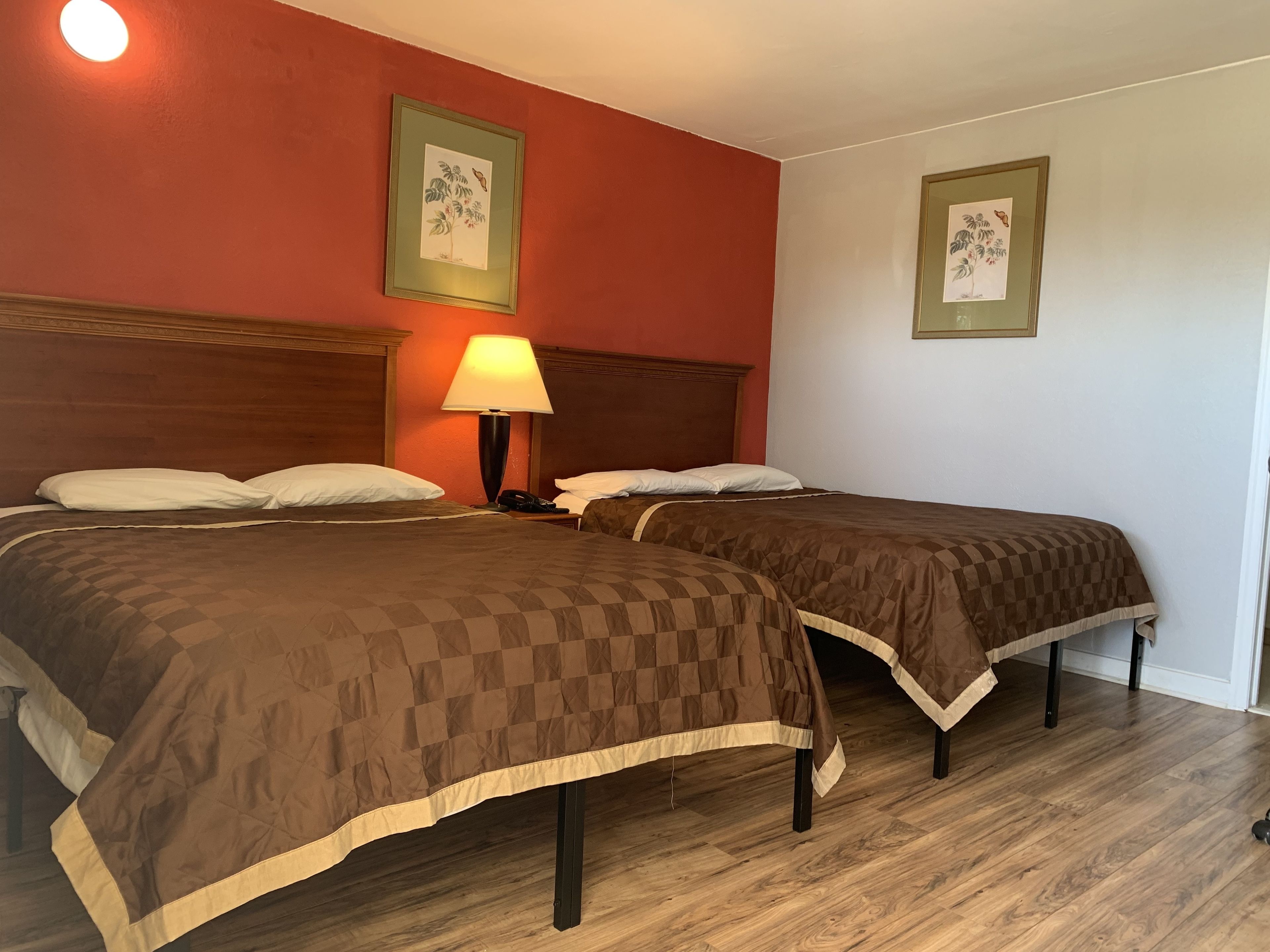 A hotel room with two beds, a nightstand with a lamp, and two framed botanical prints on the walls.