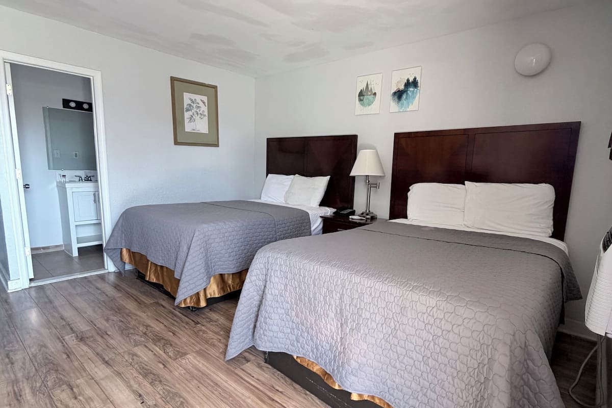 A hotel room with two beds, a wooden floor, a bathroom with a mirror, and a small desk.
