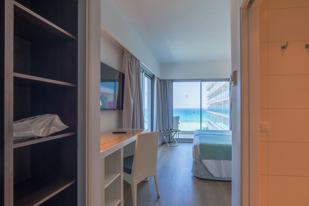 A hotel room with a desk, chair, bed, and a view of the ocean.