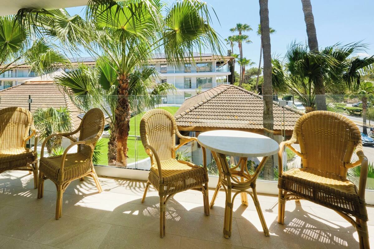 A balcony with wicker furniture, palm trees, and a view of a building with a tiled roof.