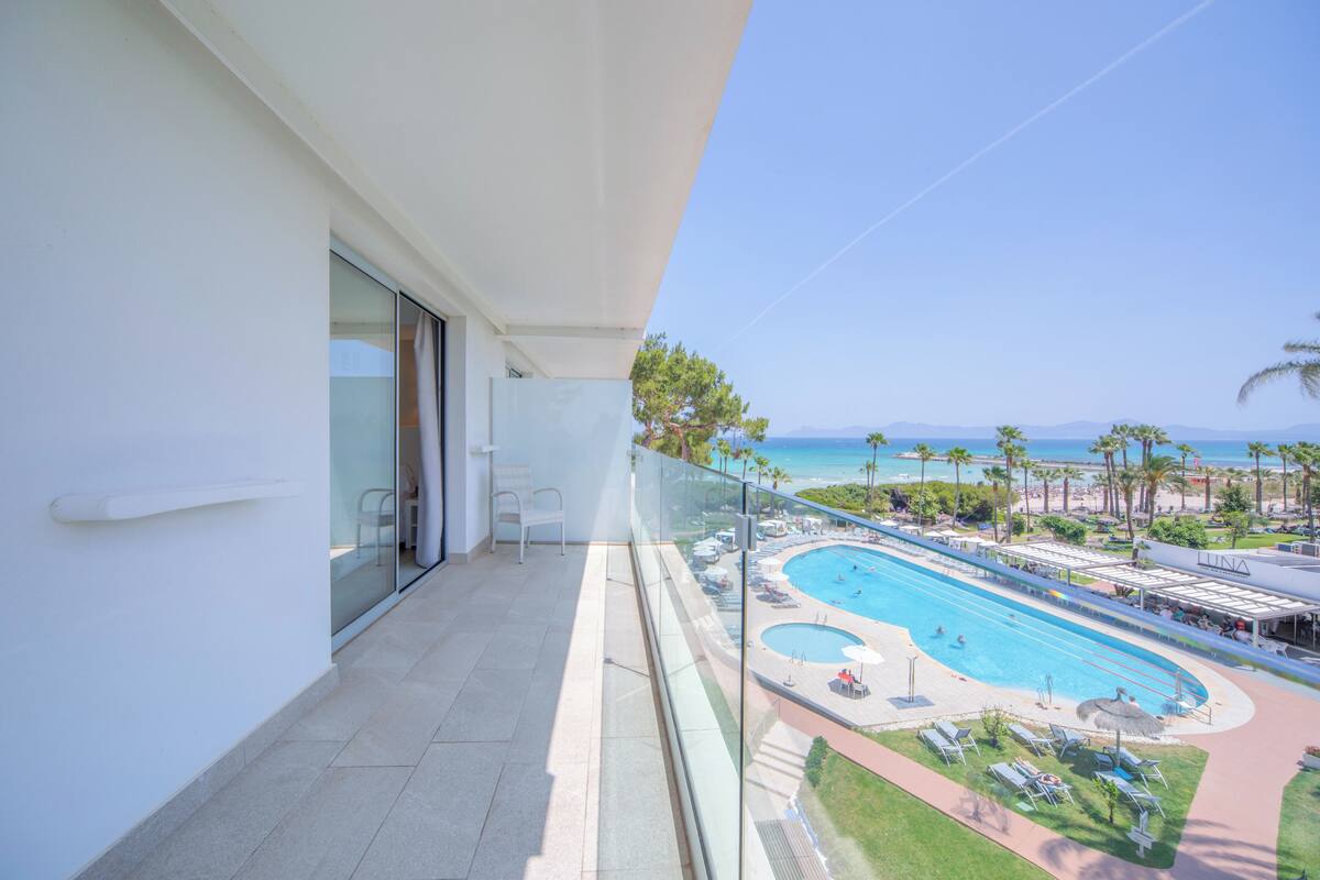 A balcony with a glass railing overlooking a swimming pool and beach area.