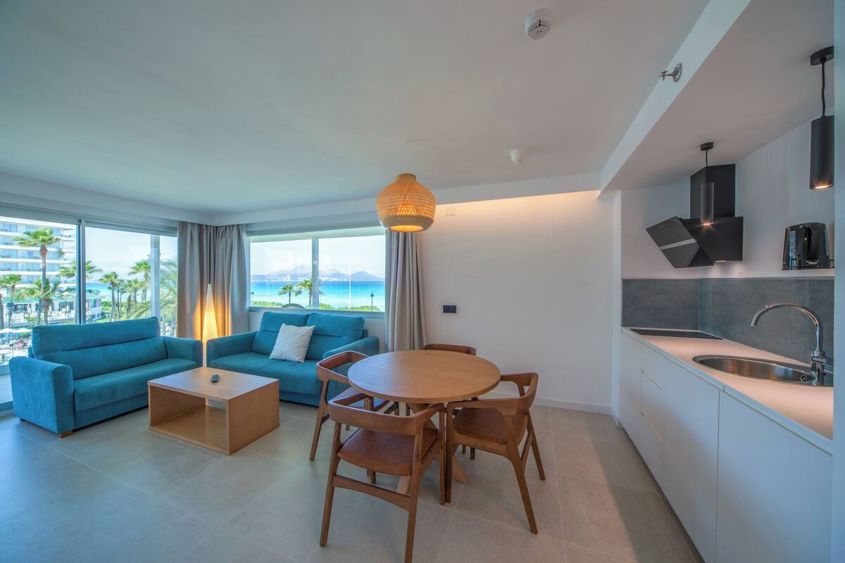 A modern living room with a sofa, coffee table, and dining table. There is a kitchen area with a sink and cabinets. Large windows offer a view of the ocean and palm trees.