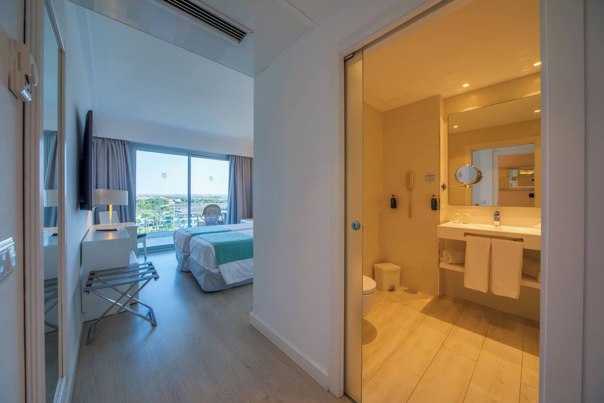 A modern hotel room with a bed, a desk with a lamp, a window with curtains, and a bathroom with a sink and mirror.