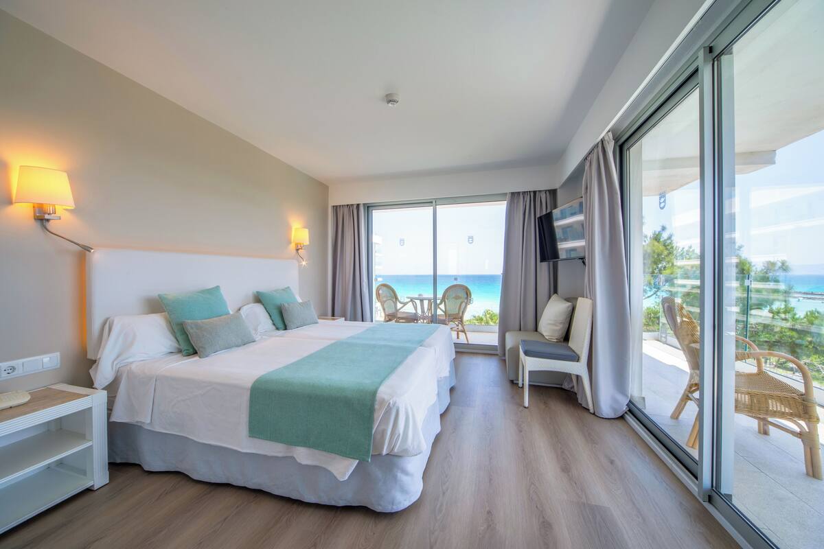 A hotel room with a large bed, a TV, a balcony with a view of the ocean, and a desk with a lamp.