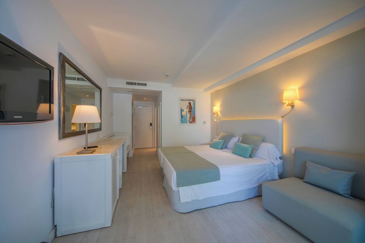 A hotel room with a bed, a sofa, a desk with a lamp, a mirror, and a wall-mounted light fixture.