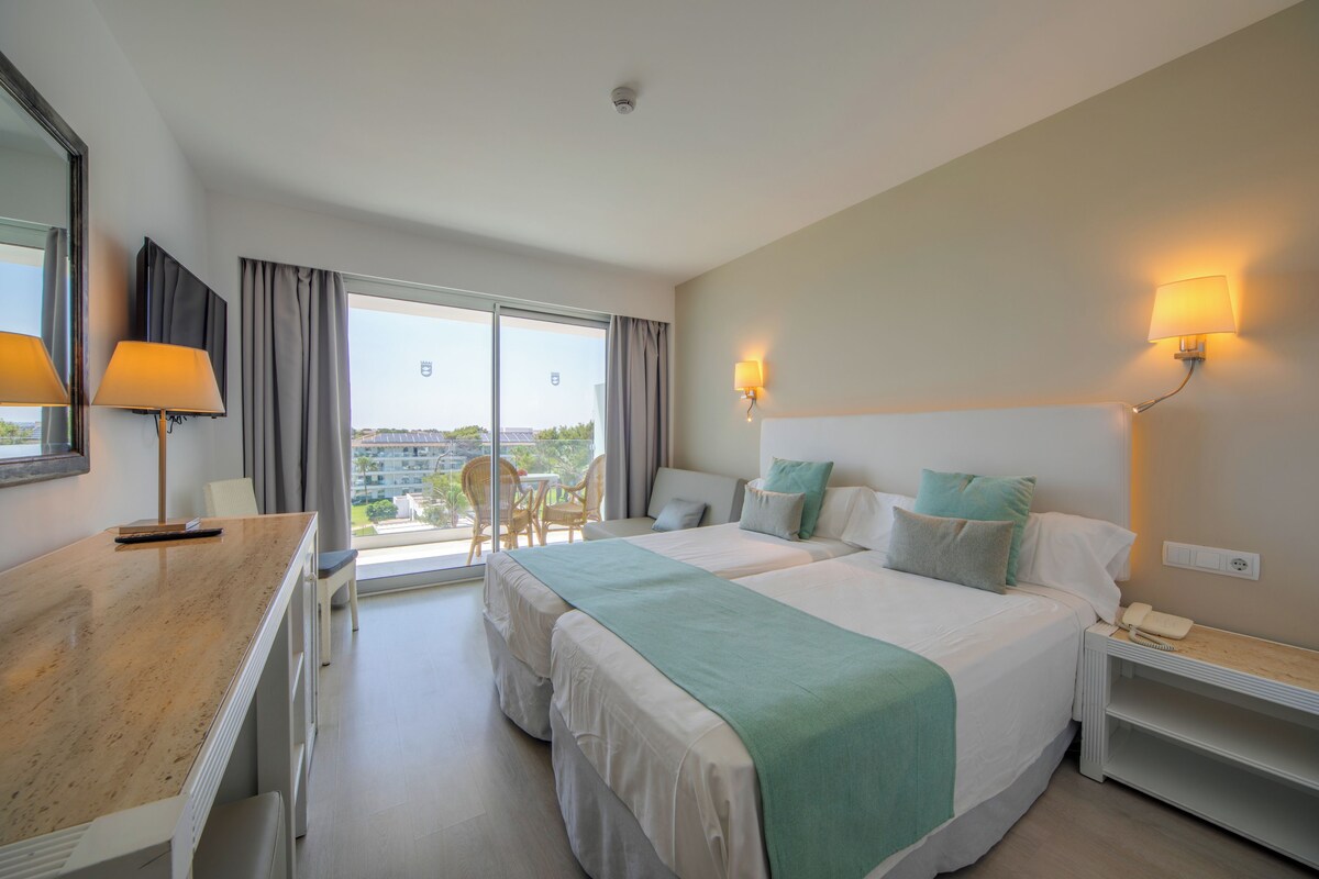 A hotel room with a bed, bedside table, lamp, and a view of the outdoors.