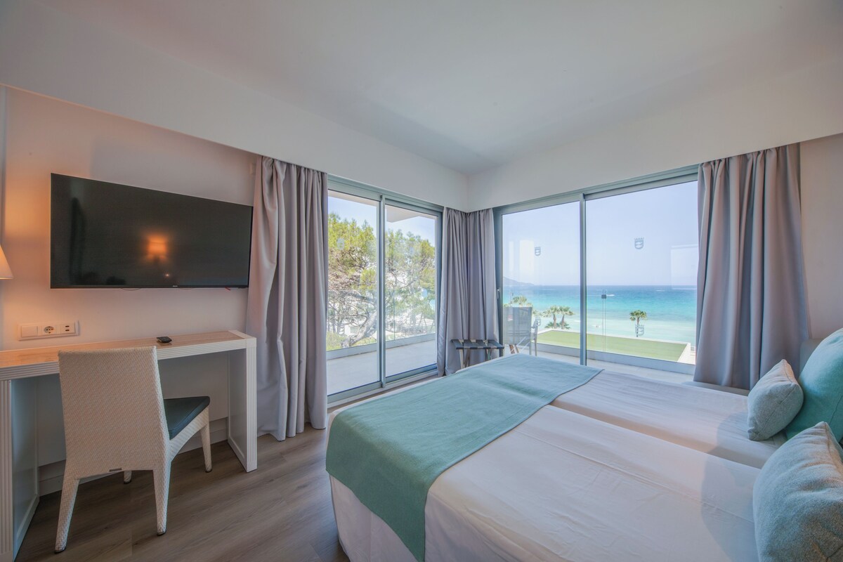 A hotel room with a bed, a desk, a chair, a TV, and large windows offering a view of the ocean.