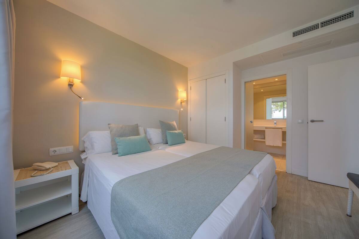 A bedroom with a bed, bedside tables, a nightstand, a mirror, and a bathroom visible through an open door.