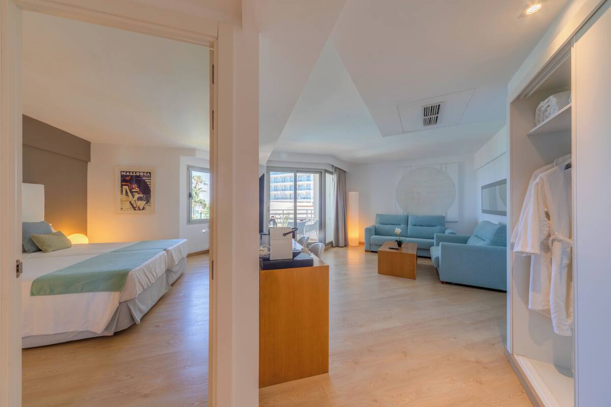 A modern hotel room with a bed, a seating area with a sofa and armchairs, a wooden coffee table, and a wardrobe with hanging clothes.