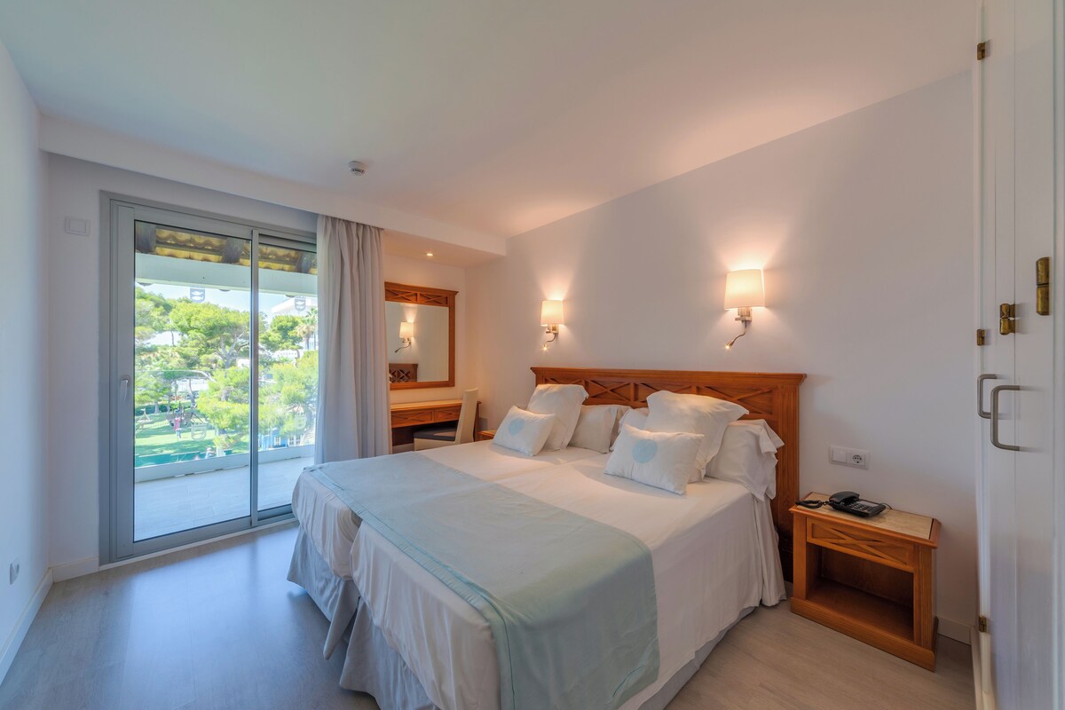 A bedroom with a large bed, a wooden headboard, two bedside tables, a nightstand, a mirror, and a sliding glass door leading to a balcony.