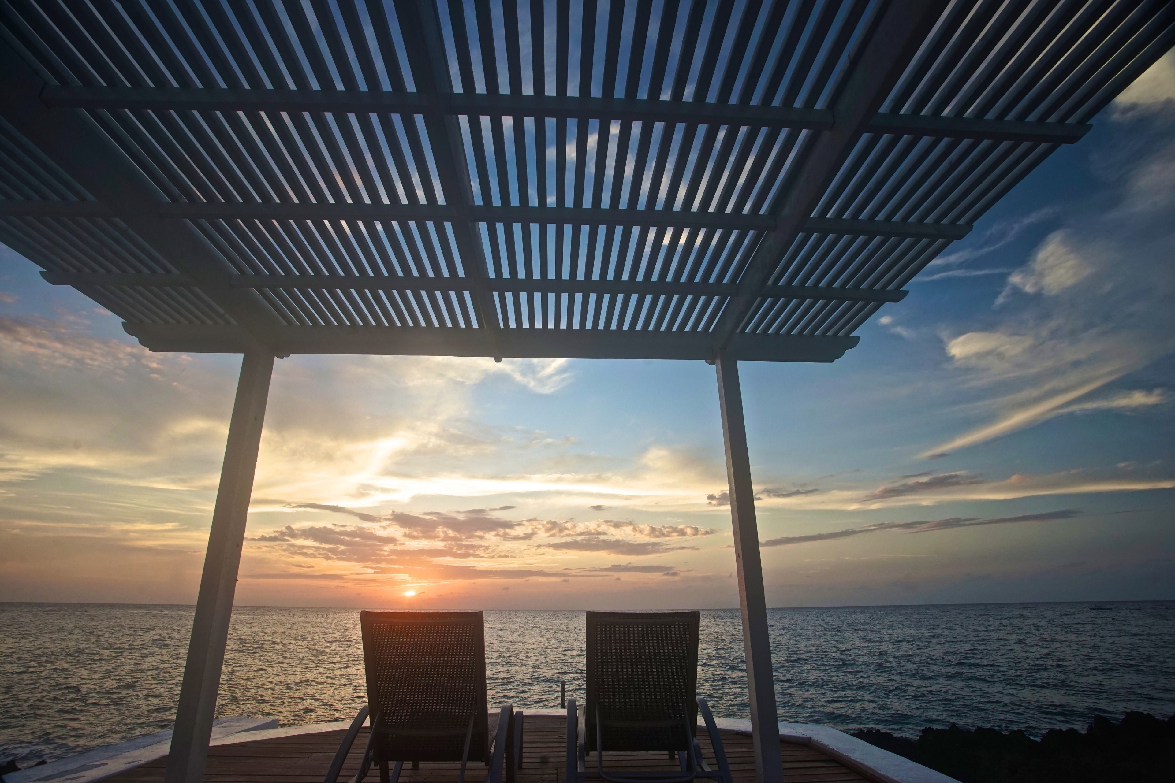 A covered outdoor seating area with two chairs facing the sea at sunset.