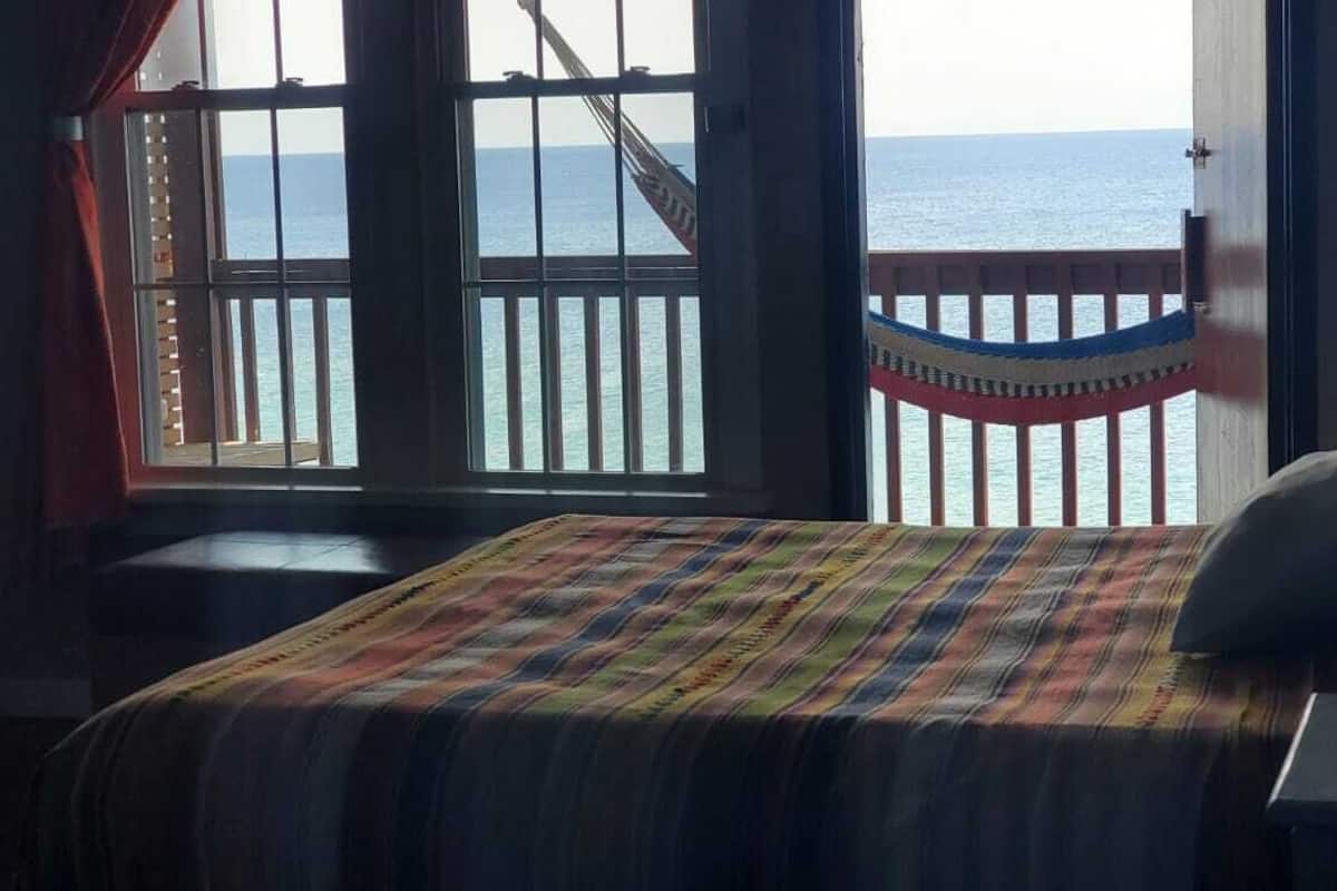 A room with a bed, a hammock, and a view of the sea through a window with a railing.